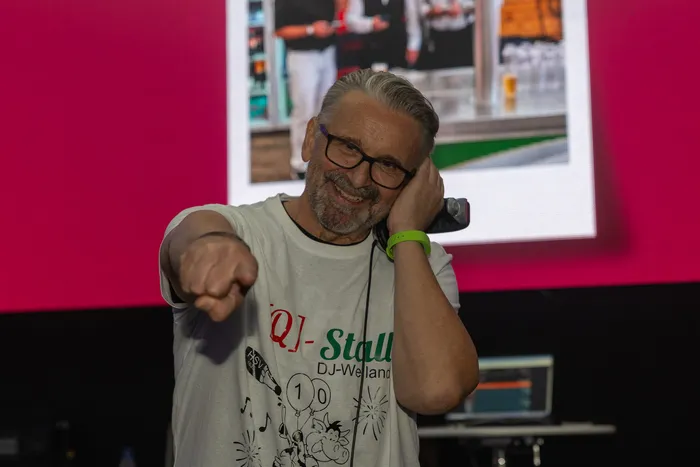 Ein lächelnder Mann mit Brille zeigt mit dem Finger in die Kamera und hält ein Telefon an sein Ohr. Er trägt ein weißes T-Shirt mit der Aufschrift „Q]-Stall DJ-Weiland“.