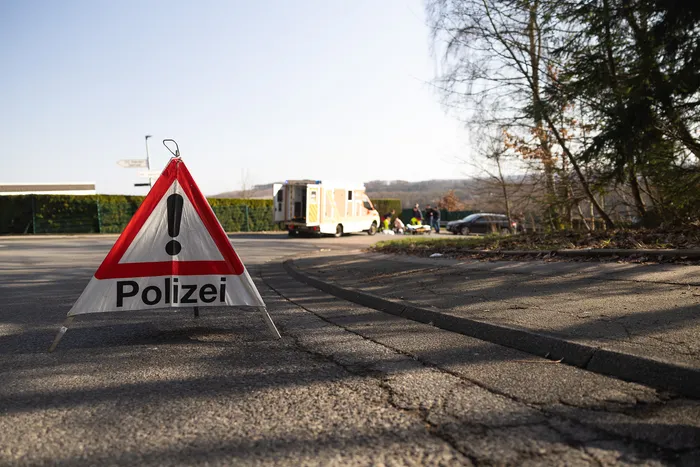 Dreieckiges Warnschild mit Ausrufungszeichen und dem Wort „Polizei“ steht am Straßenrand, im Hintergrund sind Einsatzfahrzeuge und Personen zu sehen.