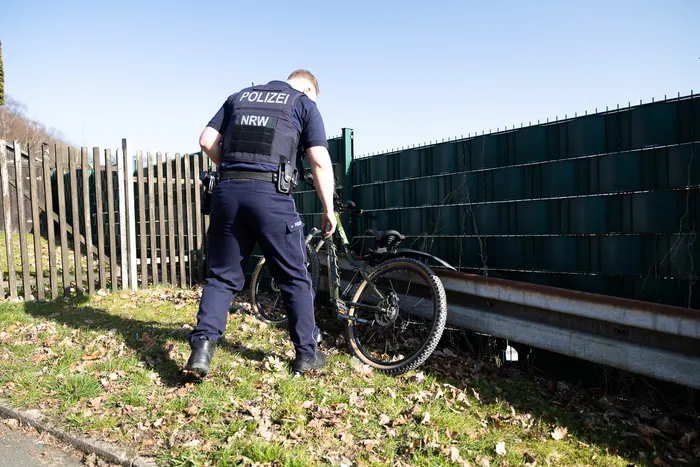 Ein Polizist in Uniform untersucht ein Mountainbike, das an einem Zaun steht. Im Hintergrund ist eine Grünfläche und ein weiterer Zaun zu sehen.