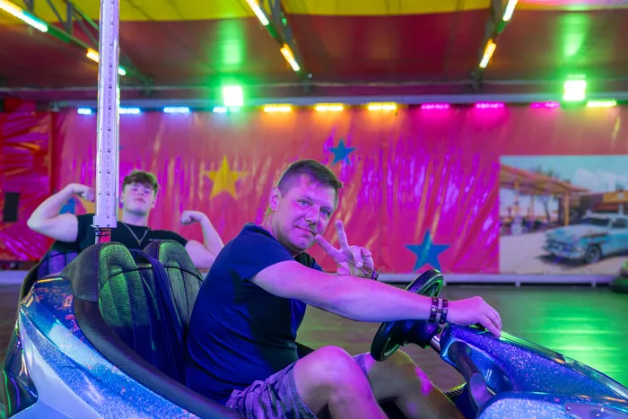 Ein Mann macht ein Peace-Zeichen in einem Bumper Car, während im Hintergrund ein junger Mann seine Muskeln anspannt.