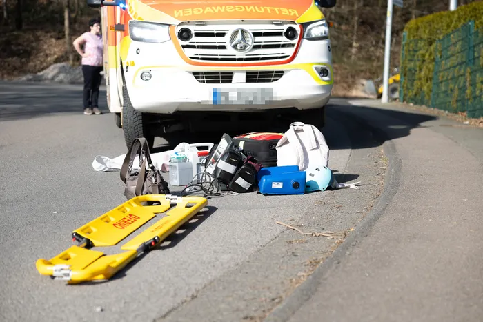 Ein Rettungswagen steht an einer Straße, um ihn herum sind medizinische Ausrüstungsgegenstände und eine Trage ausgebreitet. Im Hintergrund steht eine Person.
