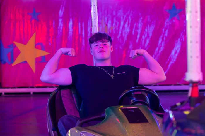 Ein junger Mann spannt seine Bizeps an, während er in einem Fahrgeschäft vor einem pinkfarbenen Hintergrund mit gelben Sternen sitzt.
