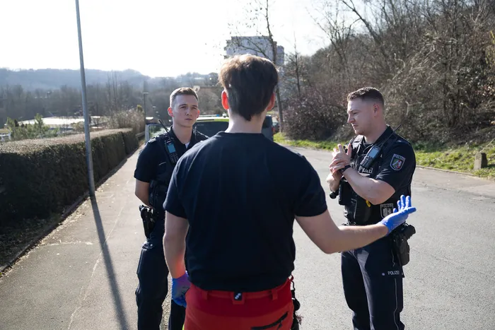 Drei Polizisten stehen auf einer Straße und sprechen mit einer Person, deren Rücken dem Betrachter zugewandt ist. Im Hintergrund sind Bäume und ein Gebäude zu sehen.
