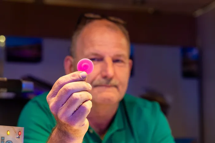 Ein Mann in einem grünen Shirt hält ein kleines, rundes, pinkfarbenes Objekt an einem Stiel in die Kamera.