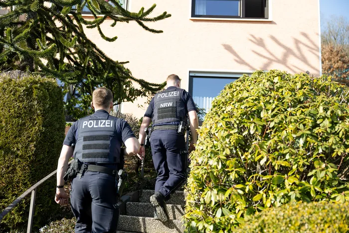 Zwei Polizisten in Uniform und Schutzwesten steigen eine Treppe zu einem Gebäude. Auf dem Rücken eines Polizisten ist „POLIZEI NRW“ zu lesen.