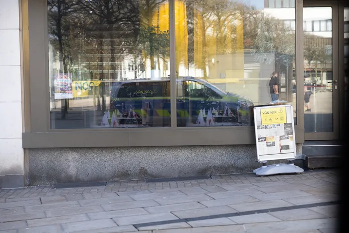 Ein helles Schaufenster reflektiert einen blauen Wagen und zeigt Dekorationen. Rechts steht ein Werbeständer mit dem Schriftzug „VAN NOO“.