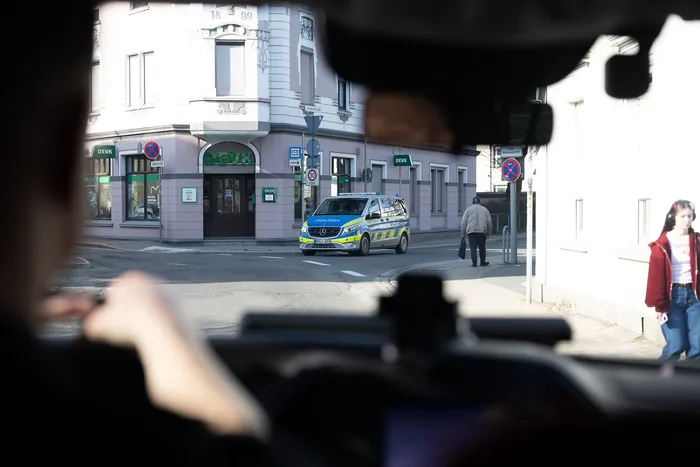 Polizeiwagen steht vor einer Bankfiliale, beobachtet von mehreren Personen mit Kameras im Vordergrund.