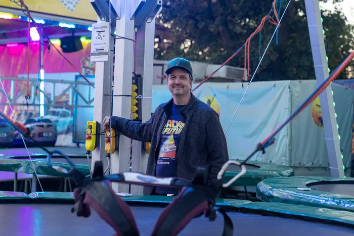 Ein lächelnder Mann mit grüner Kappe steht an einem Bedienpult einer Trampolin-Attraktion auf einer Veranstaltung.