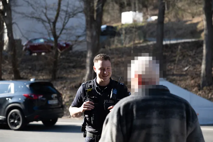 Ein Polizist spricht mit einer Person, deren Gesicht verpixelt ist, während im Hintergrund Autos und Gebäude zu sehen sind.