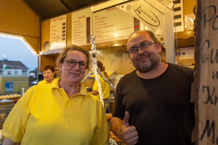 Ein Mann und eine Frau stehen vor einem Imbissstand mit Speisekarte; der Mann zeigt den Daumen nach oben.
