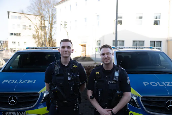 Zwei Polizisten in Uniform stehen vor zwei Streifenwagen. Im Hintergrund ein Wohngebäude.