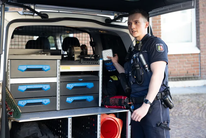 Ein Polizist in Uniform steht neben einem geöffneten Transporter mit Werkzeugausstattung und erklärt etwas mit der Hand.
