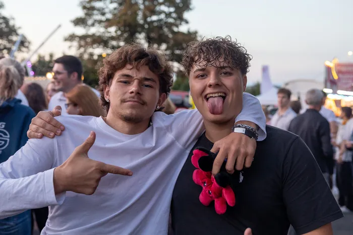 Zwei junge Männer posieren bei einer Party; einer macht eine Handgeste, der andere streckt die Zunge heraus.