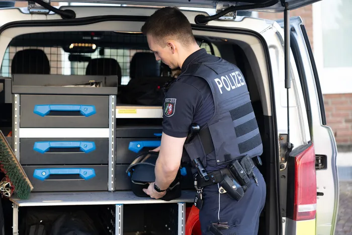 Ein Polizist in Uniform steht mit dem Rücken zum Betrachter vor einem geöffneten Transporter und nimmt Gegenstände aus einem blauen Schrank.