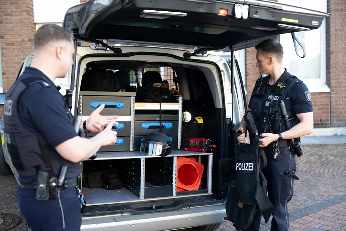 Zwei Polizisten stehen vor einem geöffneten Transporter, dessen Ladefläche mit Ausrüstung gefüllt ist. Im Inneren sind Schränke mit Werkzeug, Schutzkleidung und anderer Ausrüstung zu sehen.