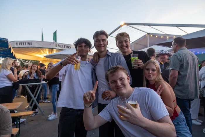 Eine Gruppe junger Menschen lacht und hält Getränke bei einer Party unter einem Festzelt.
