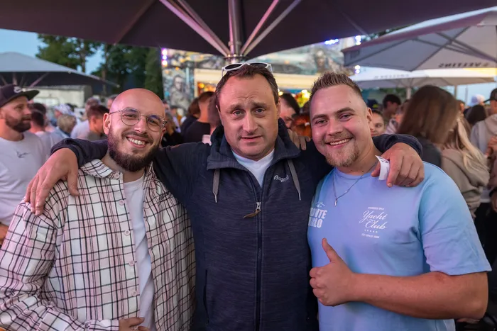 Drei lächelnde Männer posieren eng umschlungen bei der zehnten Q-Stall-Party in Schalksmühle.
