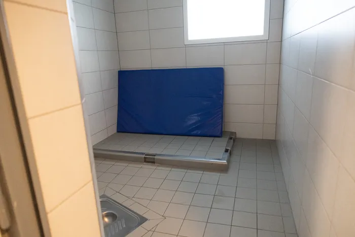 Ein karger Raum mit Fliesenwänden und -boden, in dem eine blaue Matte und eine metallene Vorrichtung liegen.