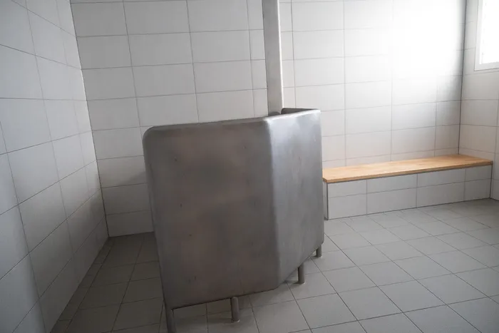 Ein metallener Verhörraum mit einer Bank an der Wand in einem hell gefliesten Raum.