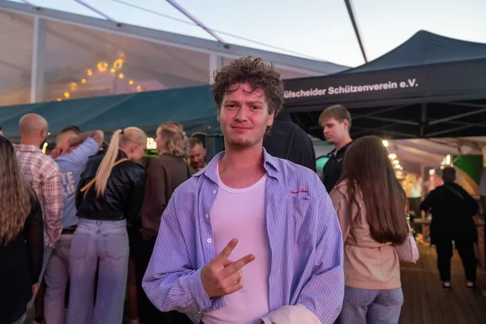 Ein junger Mann mit hellblau gestreiftem Hemd macht ein Peace-Zeichen vor einer Menschenmenge bei der Q-Stall-Party in Schalksmühle.
