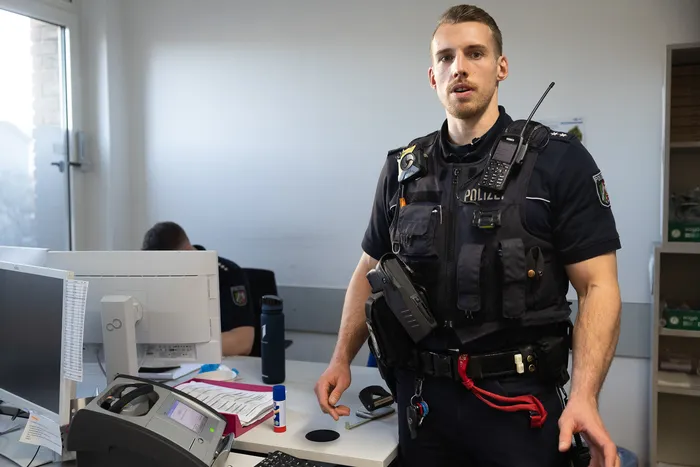 Ein Polizist in voller Ausrüstung steht in einem Büro. Er trägt eine Schutzweste mit der Aufschrift „POLIZEI“ und hat eine Pistole am Gürtel.