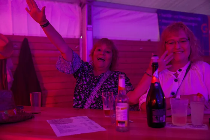 Zwei lachende Frauen sitzen bei einer Party mit pinker Beleuchtung an einem Tisch mit Getränken; eine Frau winkt mit erhobenem Arm.