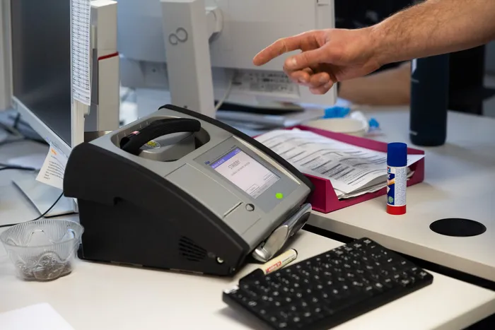 Ein Gerät zur Fingerabdruckanalyse steht auf einem Schreibtisch, während eine Person mit dem Finger darauf zeigt. Im Hintergrund sind weitere Geräte und Dokumente zu sehen.