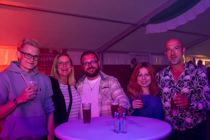 Fünf lachende Personen halten Getränke bei einer Party unter farbiger Beleuchtung.
