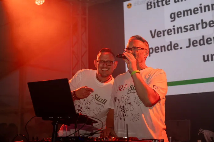 Zwei Männer in weißen T-Shirts mit Q-Stall-Logo stehen an einem DJ-Pult, einer hält ein Mikrofon.