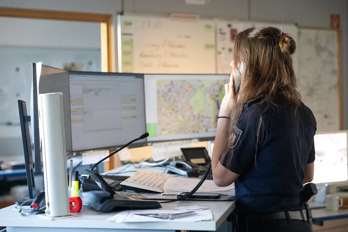 Eine Polizistin mit zurückgebundenen Haaren sitzt an einem Schreibtisch vor zwei Computerbildschirmen und telefoniert. Auf dem Schreibtisch befinden sich ein Headset, ein Telefon und verschiedene Arbeitsmaterialien.