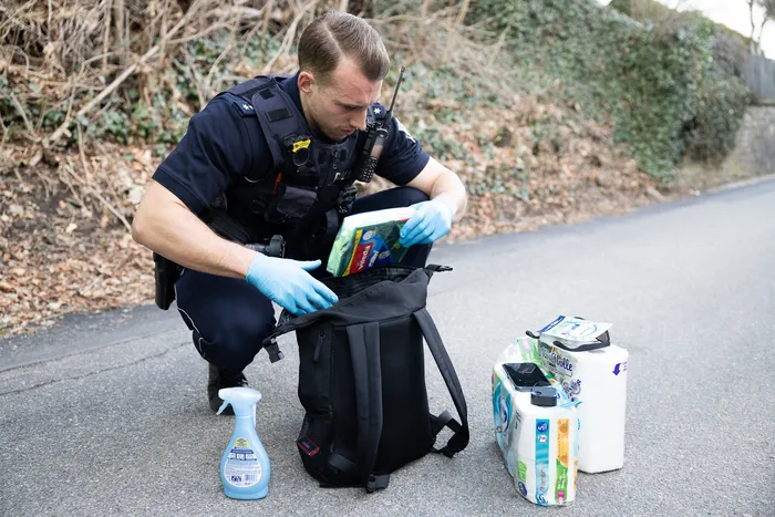 Ein Polizist in Uniform und blauen Handschuhen durchsucht eine schwarze Tasche am Straßenrand. Neben der Tasche stehen Reinigungsmittel und Babypflegeprodukte.