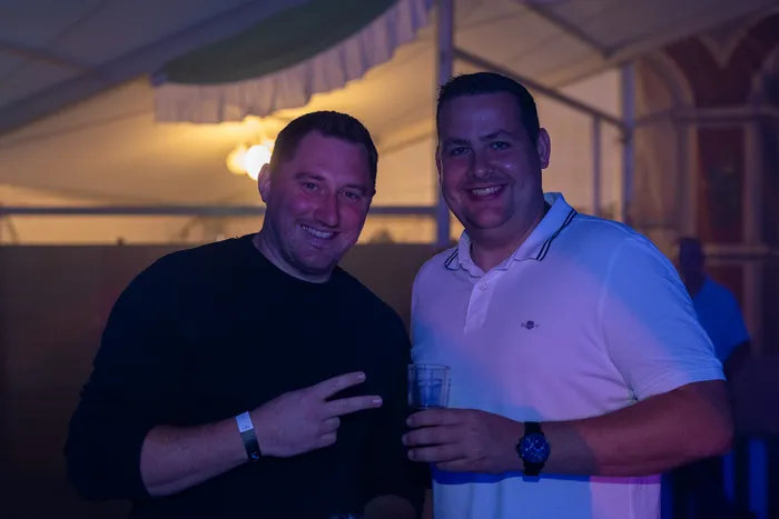 Zwei lächelnde Männer bei einer Party; einer trägt ein schwarzes Oberteil und macht ein Peace-Zeichen, der andere ein weißes Poloshirt und hält ein Glas.