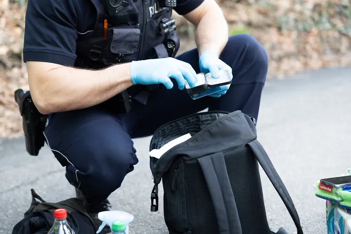 Ein Polizist in Uniform und blauen Handschuhen untersucht einen kleinen, dunklen Gegenstand über einem geöffneten Rucksack.