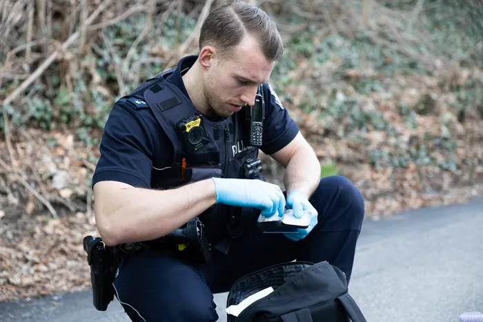 Ein Polizist in Uniform hockt auf der Straße und verpackt Beweismittel in einer Plastiktüte. Er trägt blaue Handschuhe und blickt konzentriert auf seine Arbeit.