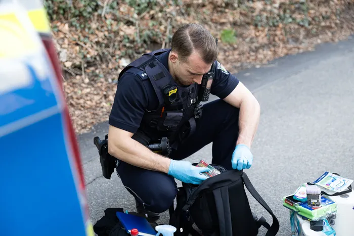 Ein Polizist in Uniform hockt auf der Straße und durchsucht einen schwarzen Rucksack. Er trägt blaue Handschuhe und hat ein Funkgerät am Gürtel.