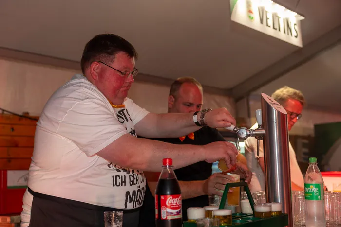 Ein Mann zapft ein Getränk aus einer Zapfanlage an einem Tresen. Im Hintergrund sind weitere Personen und ein Veltins-Schild zu sehen.