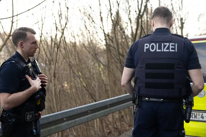 Zwei Polizisten im Einsatz, einer hält eine Waffe bereit, während der andere den Blick in die Ferne richtet. Beide tragen Schutzwesten mit der Aufschrift „POLIZEI“.