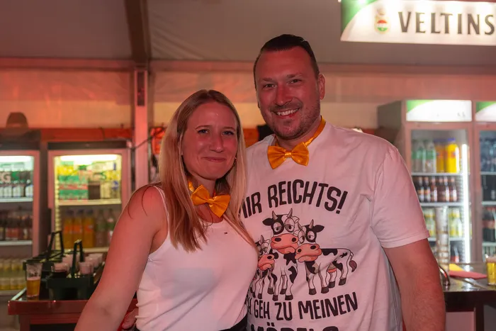 Ein lächelndes Paar mit gelben Fliegen posiert vor einer Bar; der Mann trägt ein weißes T-Shirt mit Kuh-Motiven.