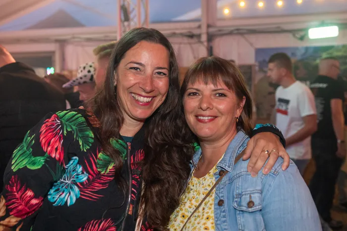 Zwei lächelnde Frauen posieren eng beieinander; eine trägt ein buntes tropisches Oberteil, die andere eine Jeansjacke.