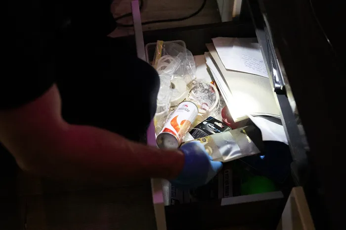 Eine Person mit Handschuhen leuchtet mit einer Taschenlampe in eine geöffnete Schublade, die mit verschiedenen Gegenständen gefüllt ist, darunter Lebensmittelverpackungen und Papier.