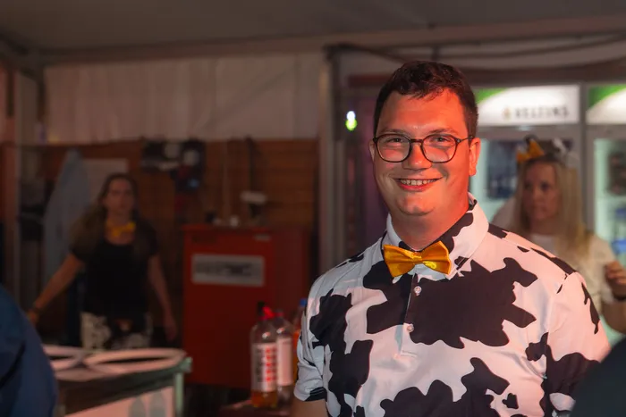 Ein lächelnder Mann mit Brille trägt ein schwarz-weißes Hemd im Kuhmuster und eine gelbe Fliege.