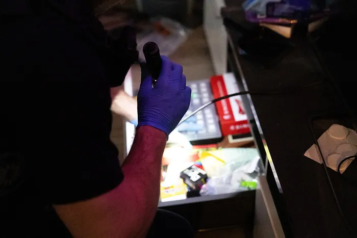Eine Hand mit blauem Handschuh hält eine kleine Plastiktüte mit Inhalt über einem Tisch mit Gegenständen und Geräten.