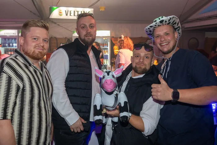 Vier Männer posieren bei der Q-Stall-Party, einer hält eine kleine rosa-weiße Kuh und ein anderer trägt einen Hut mit Kuhmuster.
