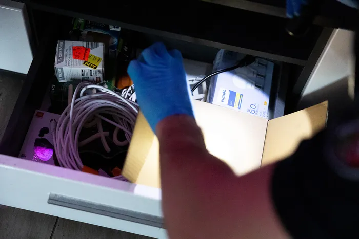 Ein Polizist leuchtet mit einer Taschenlampe in eine Schublade voller Kabel, Verpackungen und einer Schachtel mit Glühbirnen.