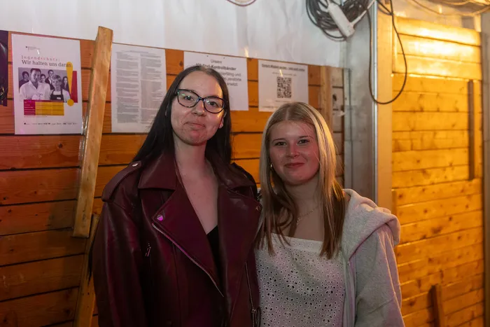 Zwei junge Frauen stehen vor einer Holzwand mit Plakaten; eine trägt eine weinrote Lederjacke und eine Brille, die andere ein weißes Oberteil.