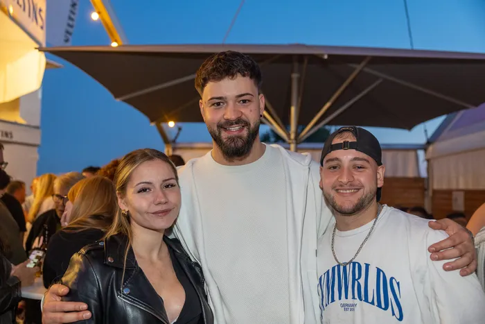 Drei lächelnde junge Menschen posieren bei der Q-Stall-Party unter einem großen Sonnenschirm.