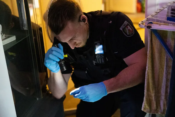 Ein Polizist in Uniform und blauen Handschuhen fotografiert mit einer Kamera einen Gegenstand am Boden in einem Raum.