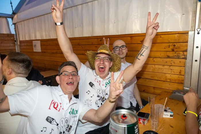 Jubelnde Gäste in weißen T-Shirts bei der Q-Stall-Party, ein Mann im Zentrum trägt einen Strohhut und macht ein Peace-Zeichen.