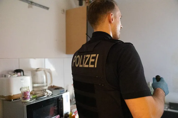 Ein Polizist mit Schutzweste und Handschuhen steht mit dem Rücken zum Betrachter in einer Küche. Auf der Weste ist deutlich „POLIZEI“ zu lesen.