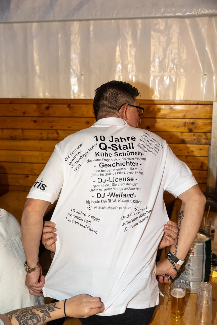 Rückansicht einer Person in einem weißen T-Shirt mit dem Aufdruck „10 Jahre Q-Stall“.
